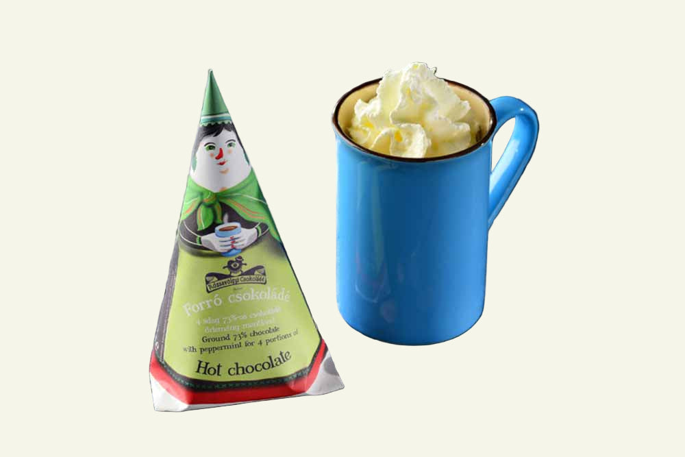 Green hot chocolate packet with a character on a wooden surface next to a blue mug with whipped cream.